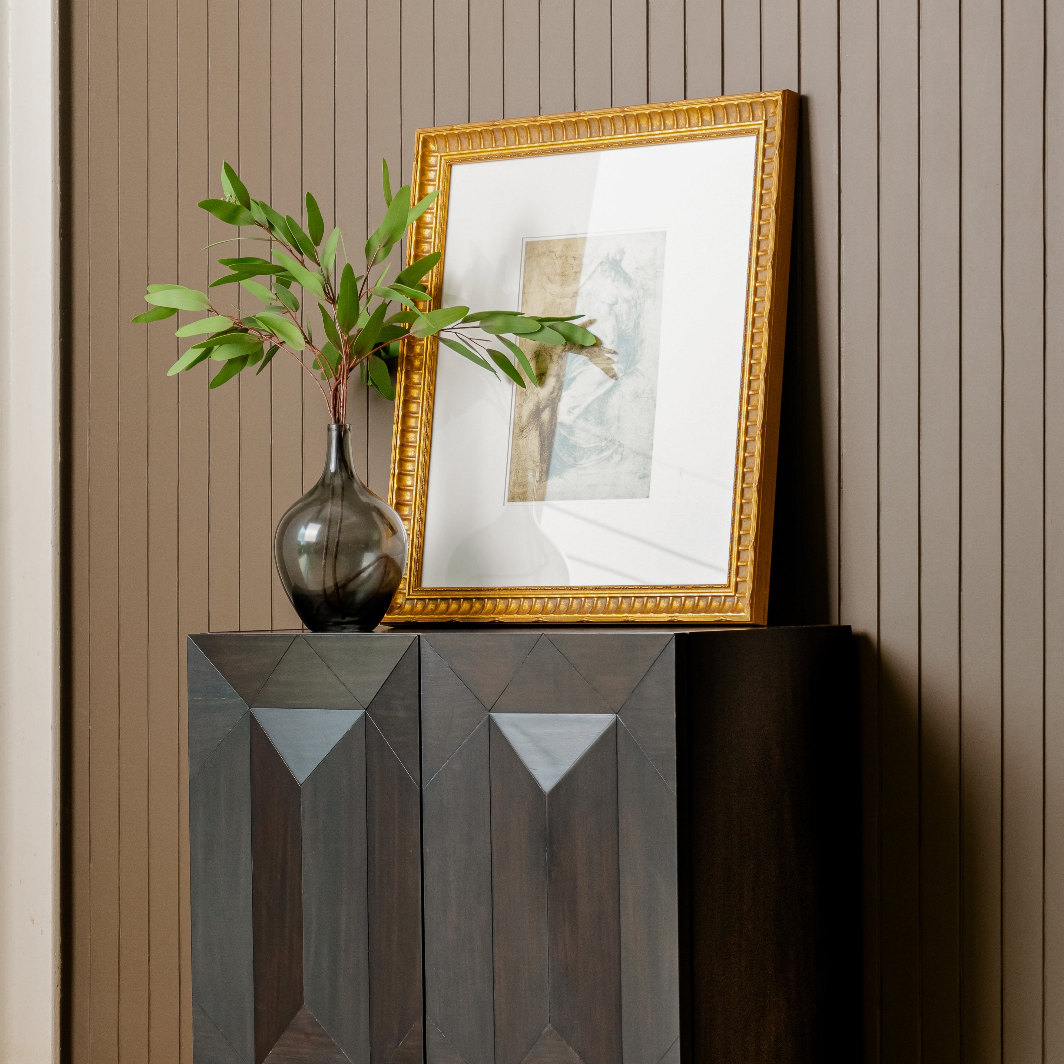 Decorative setup with a gold-framed mirror, vase with greenery, and geometric-patterned Black Averi Modular Cabinet against a striped wall.