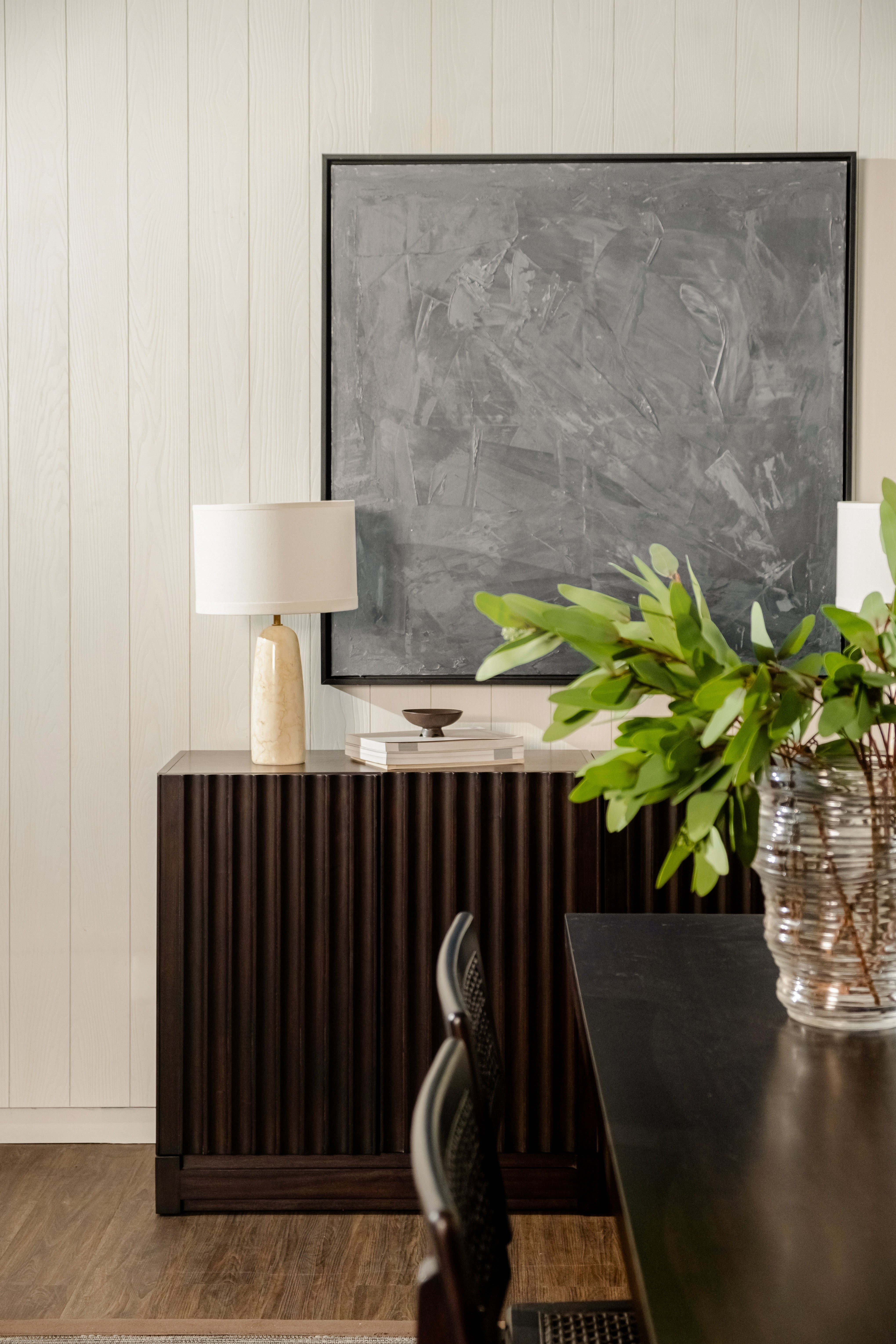 Dining room with a dark wooden console table, lamp, and abstract painting on a light wood paneled wall.