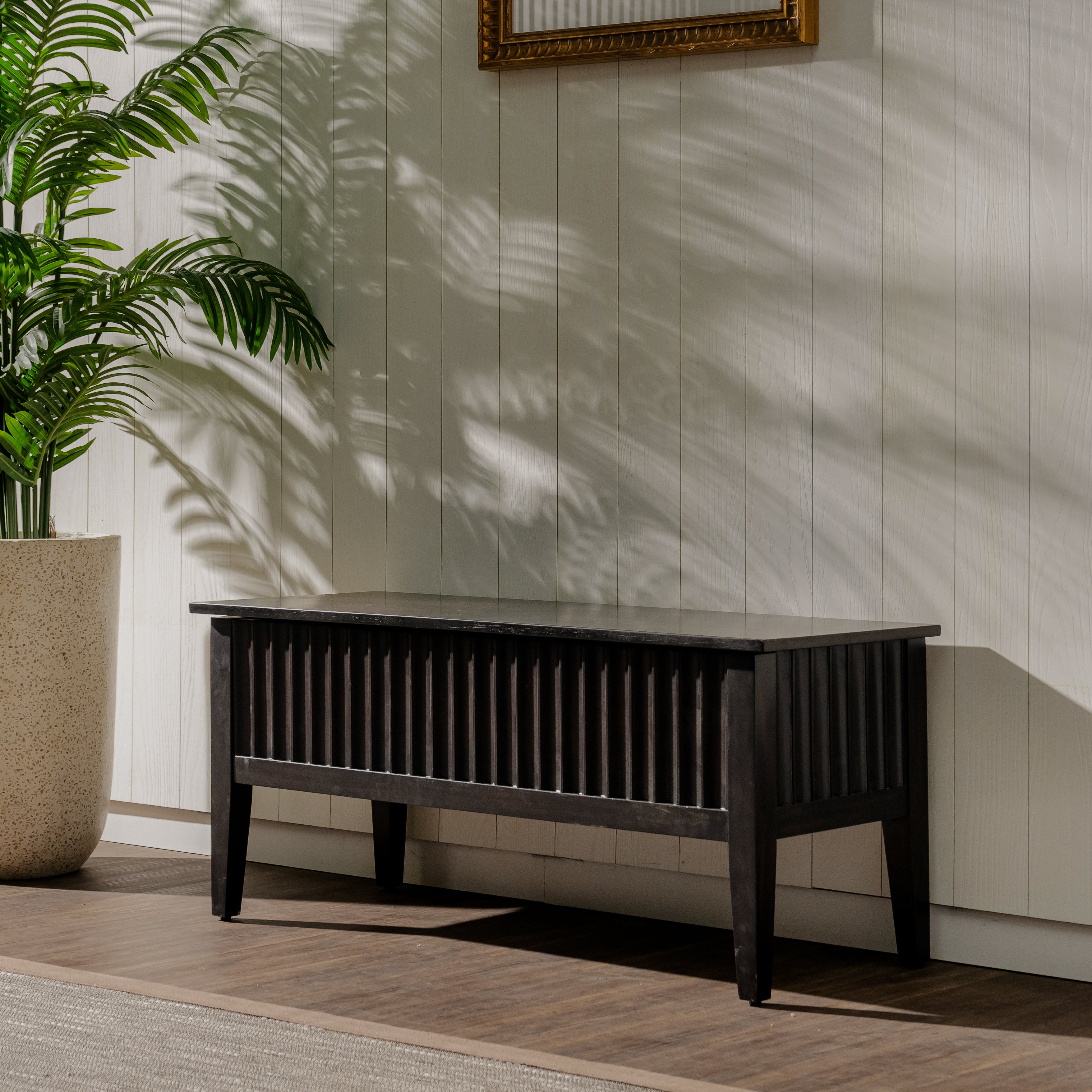 Dark wooden bench against a light paneled wall with a plant to the left.