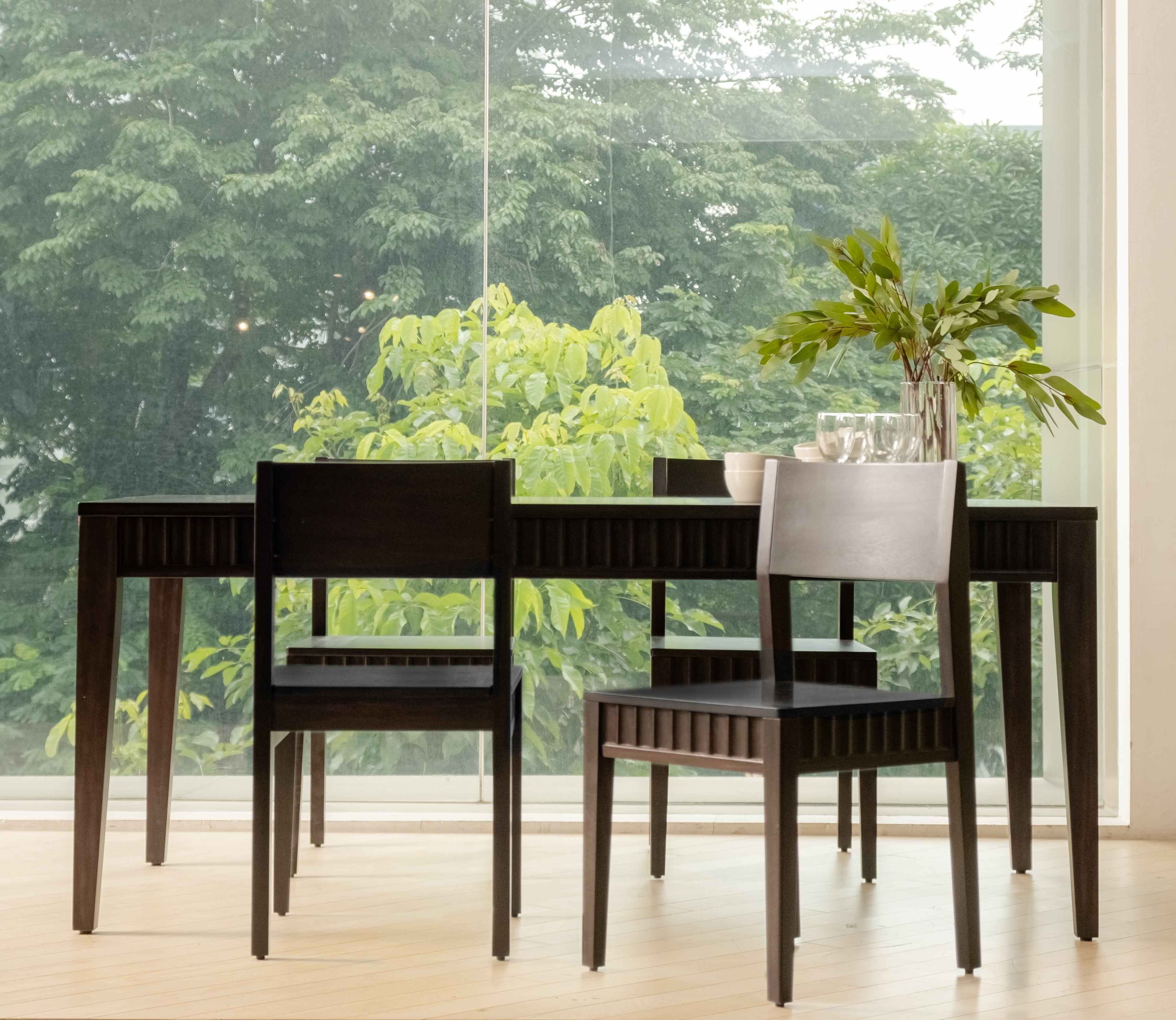 Dining table set with chairs in a room with large windows showing greenery outside.