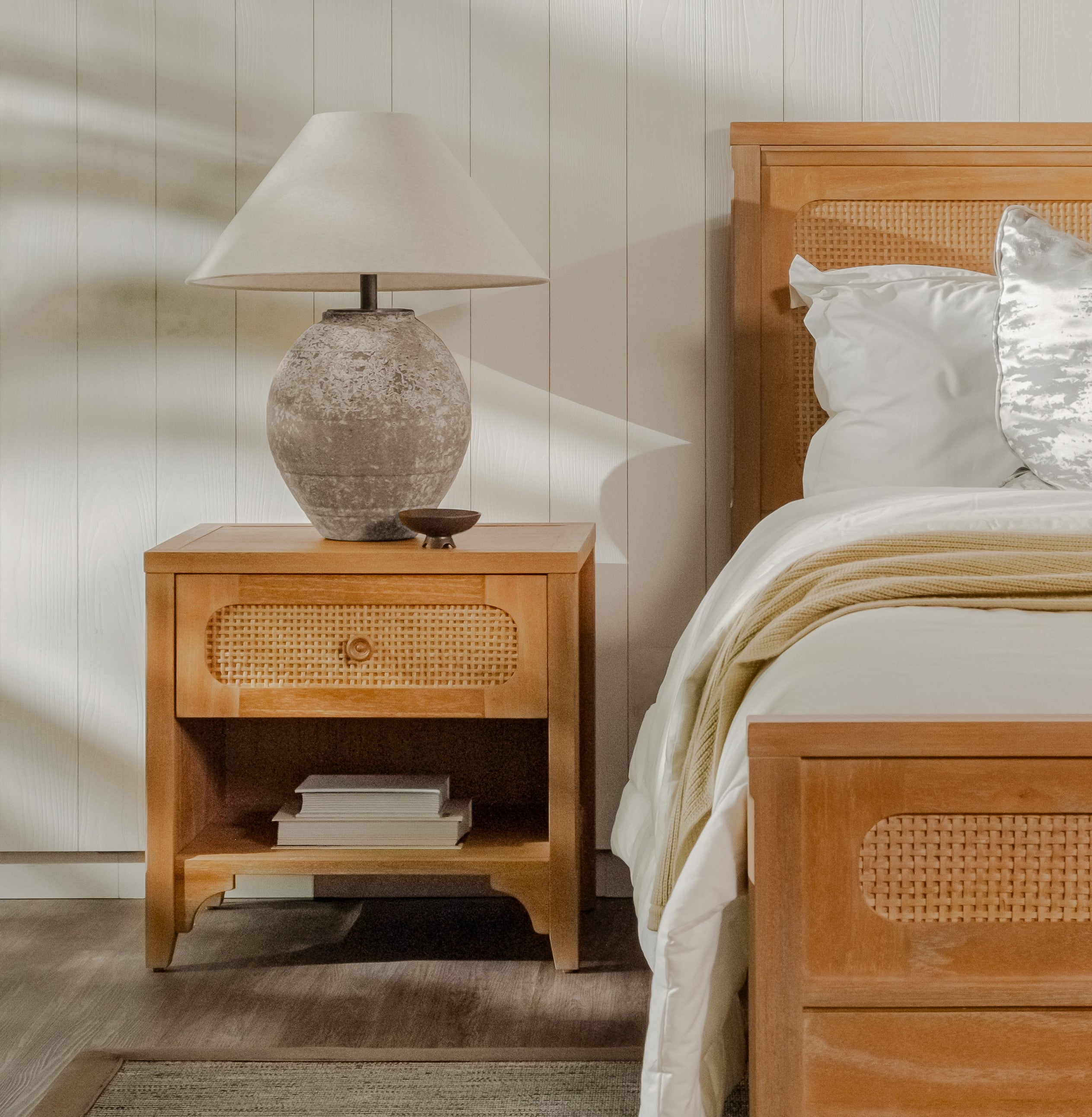 Wooden nightstand with textured lamp and bed with white bedding in a bedroom setting.
