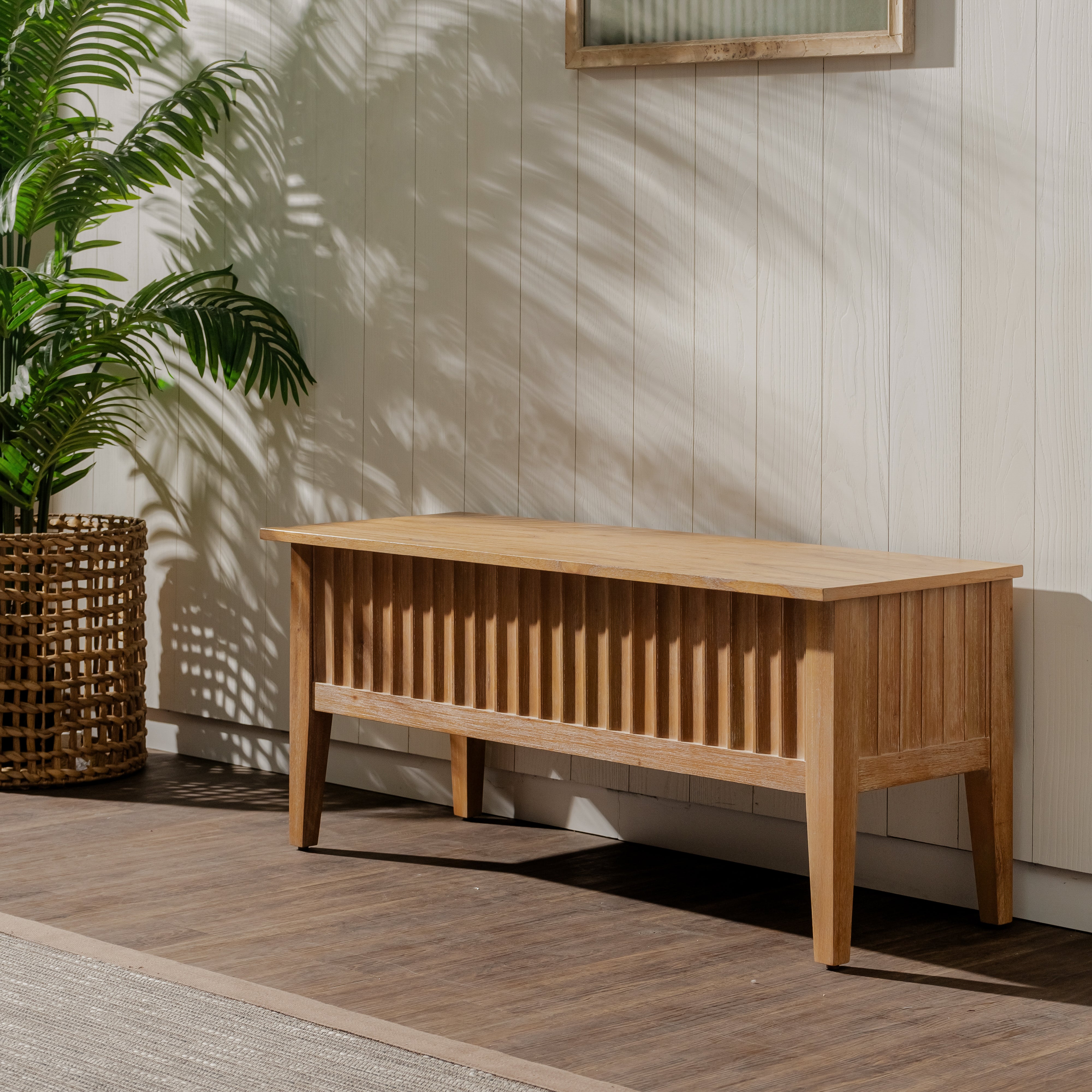 Wooden bench against a light wooden paneled wall with a plant to the left.