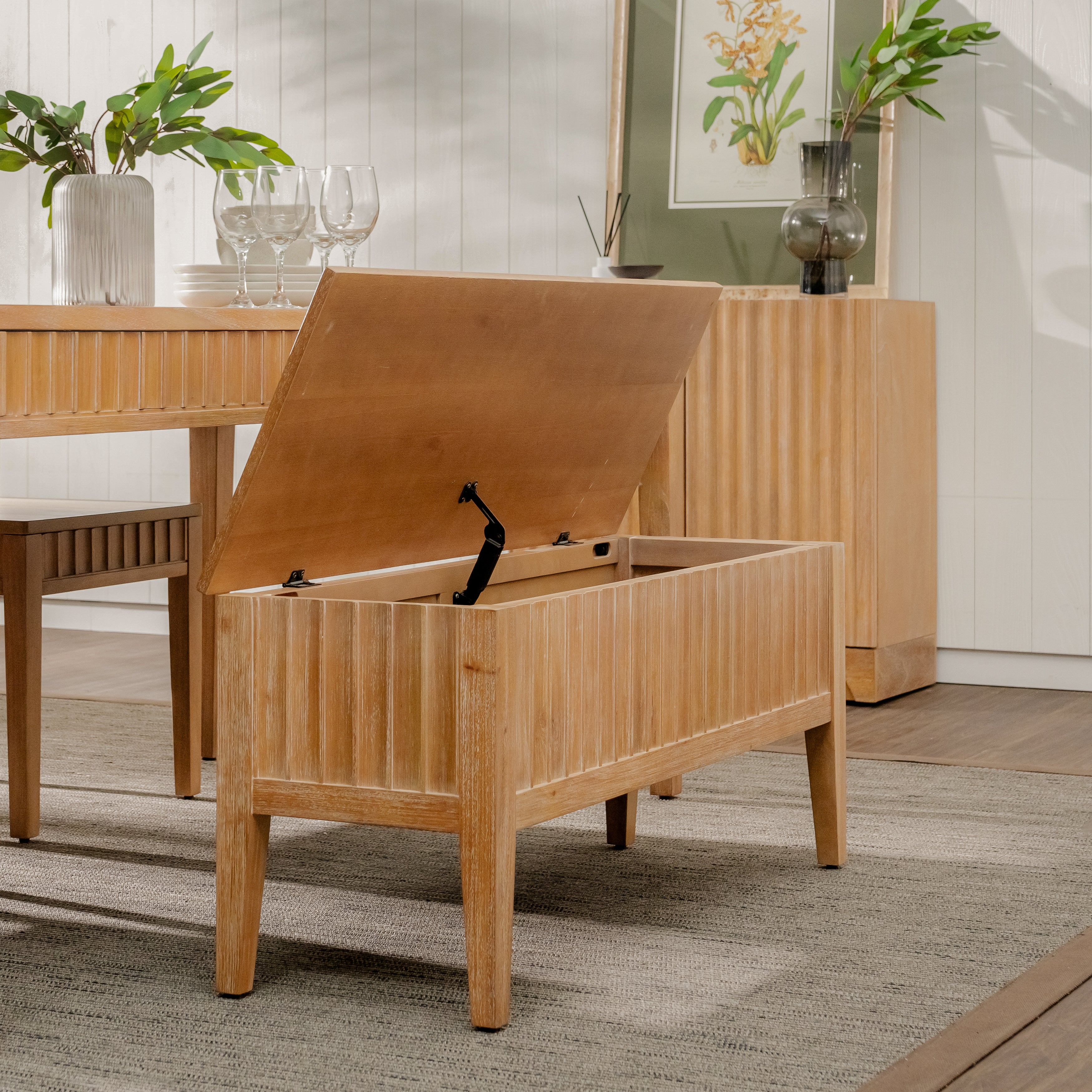 Wooden storage bench with open lid in a room with a table and plants.