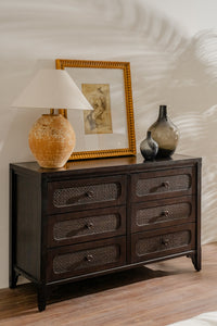 Dark wooden dresser with decorative items including a lamp, framed picture, and vases on a light wood floor.