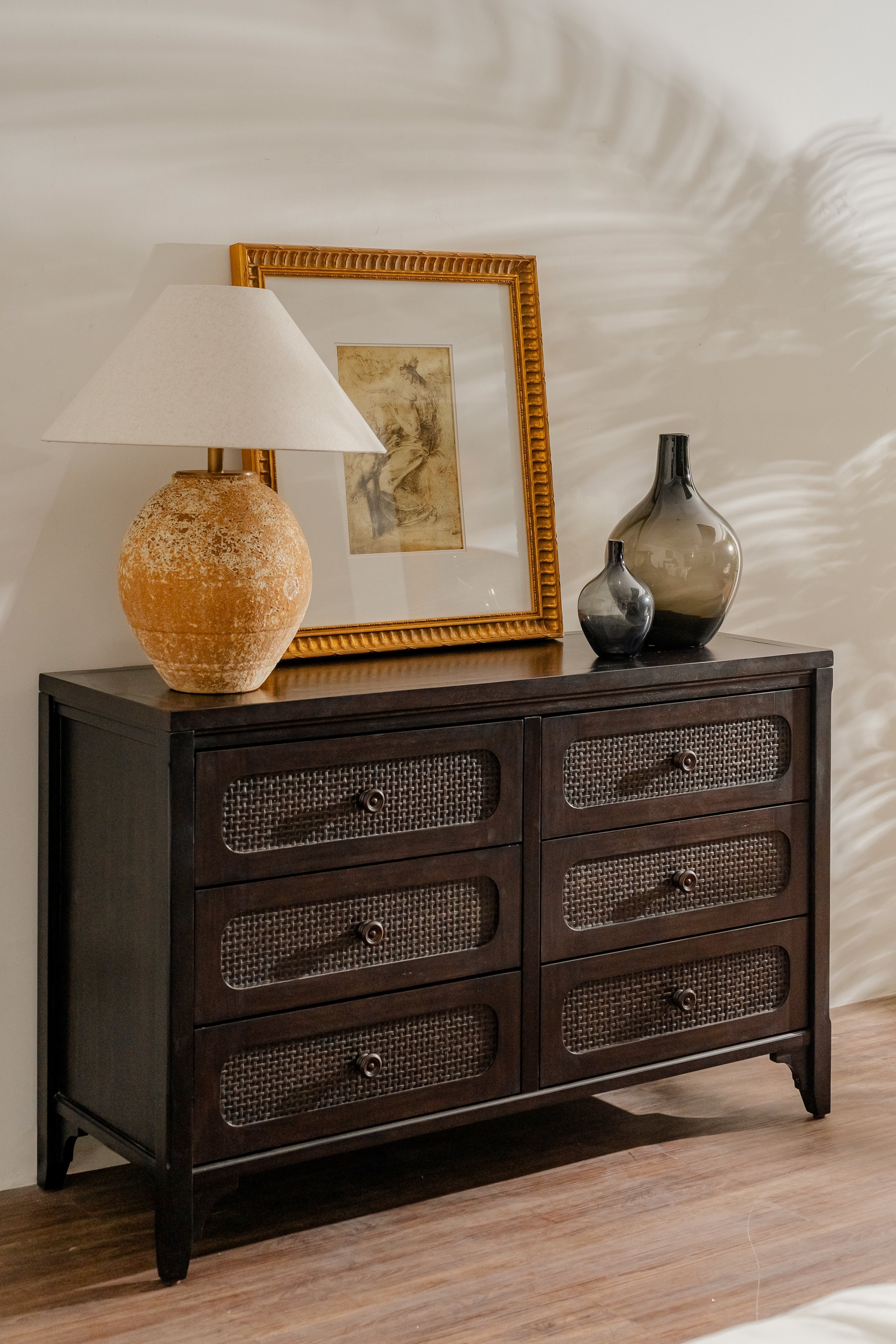 Dark wooden dresser with decorative items including a lamp, framed picture, and vases on a light wood floor.