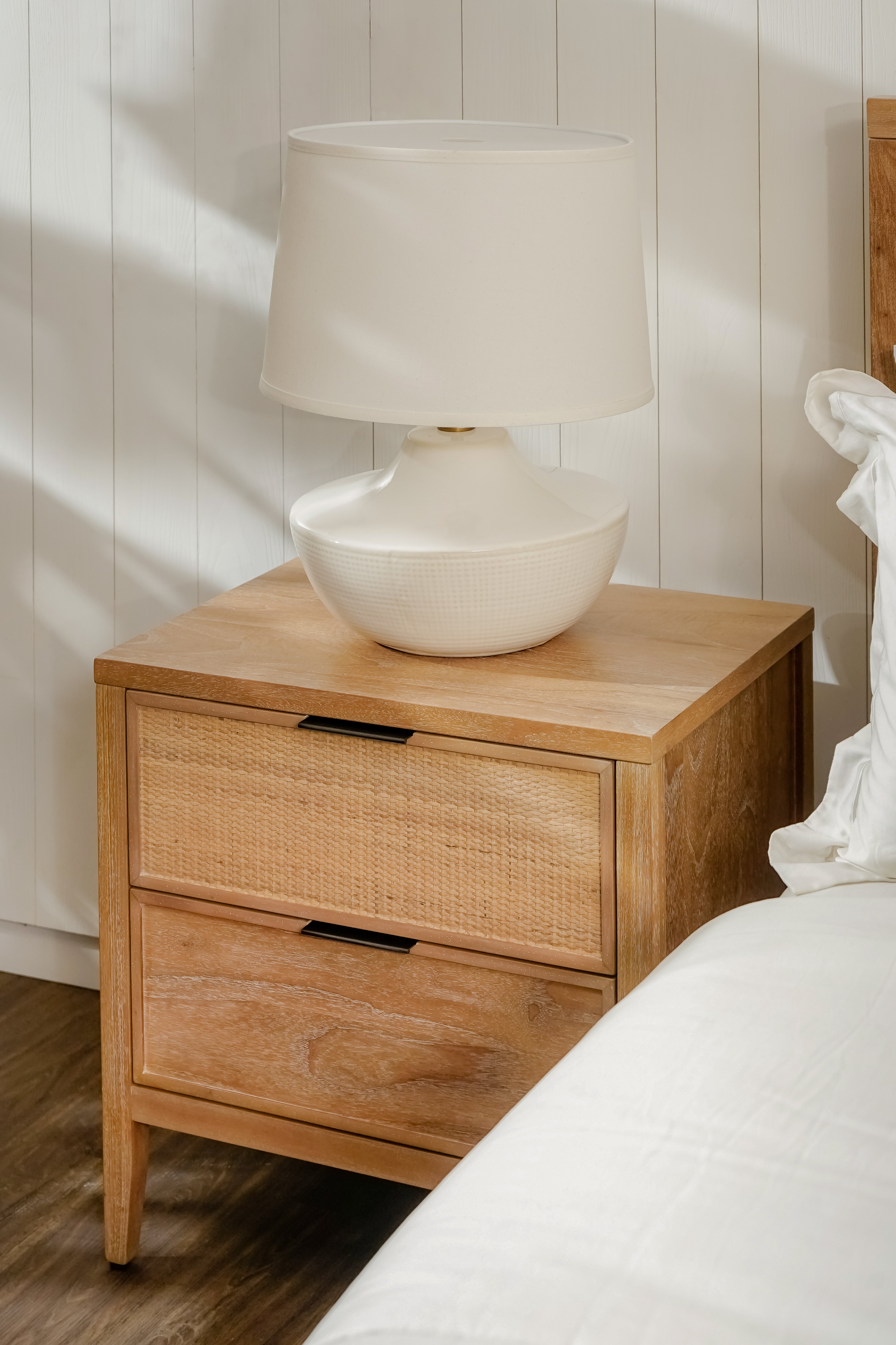 Wooden nightstand with wicker drawers and a white lamp on top against a neutral wall