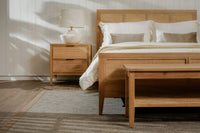 Wooden bedroom furniture set with bed, nightstand, and side table in a room with sunlight filtering through.